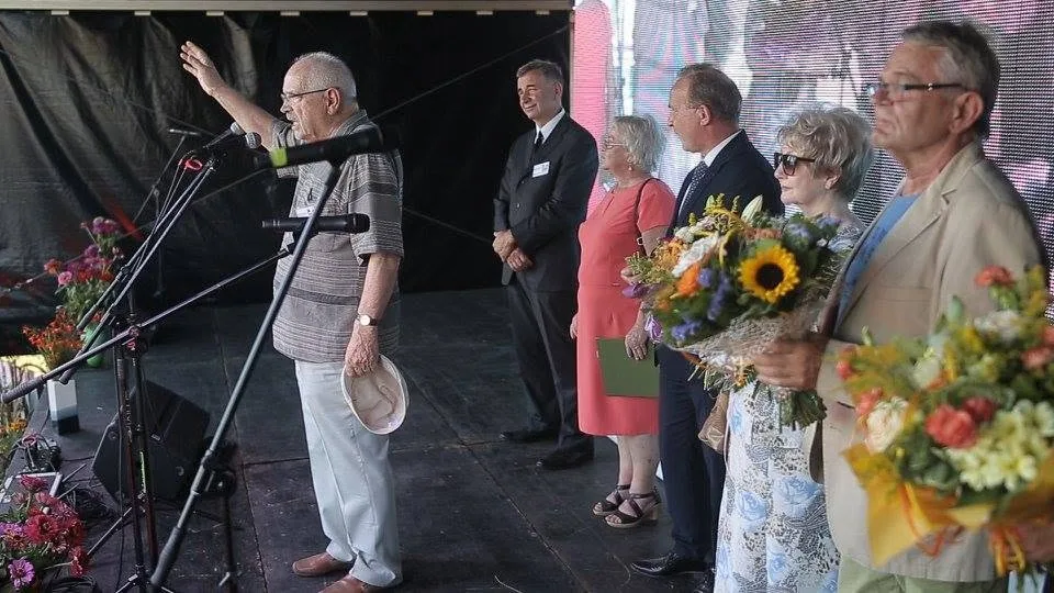 Przemówienie na scenie XIII Festynu; stojący obok goście trzymają bukiety słoneczników.